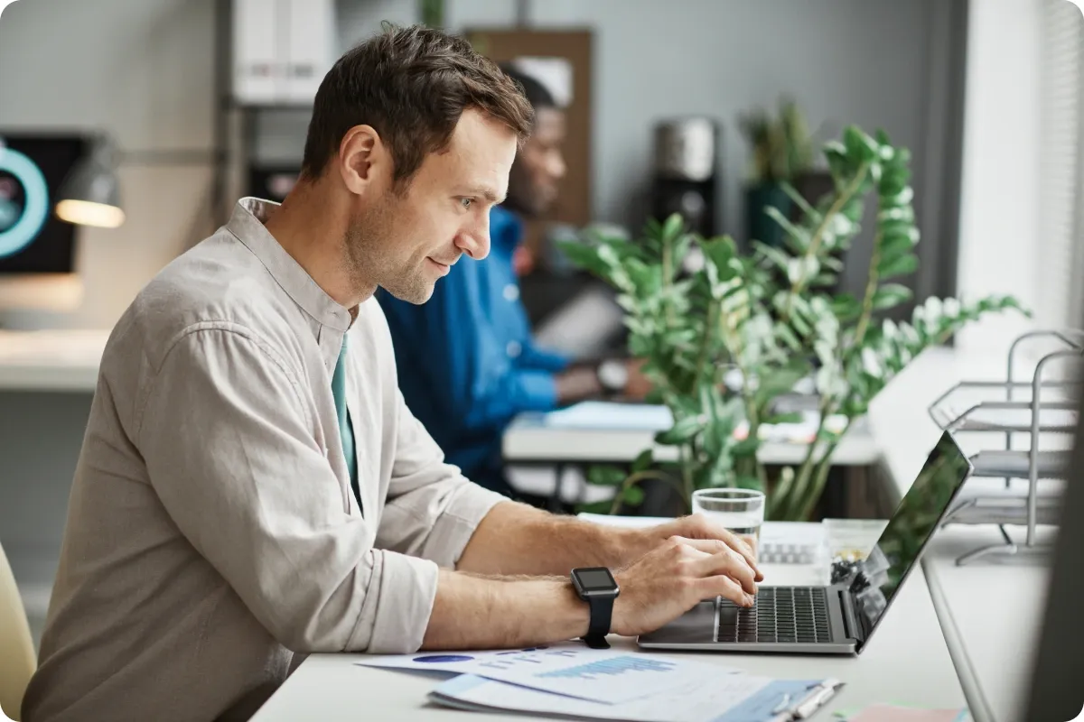 A man working at a laptop in an open-plan office, a colleague at a desk behind him